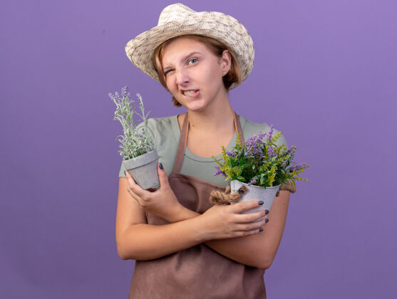 斯拉夫自信的年轻斯拉夫女园丁戴着园艺帽眨眼 手里拿着紫色花盆里的花女性园艺花
