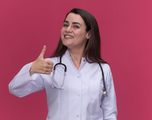 长袍面带微笑的年轻女医生穿着医用长袍 听诊器大拇指朝粉红色竖起拇指女年轻