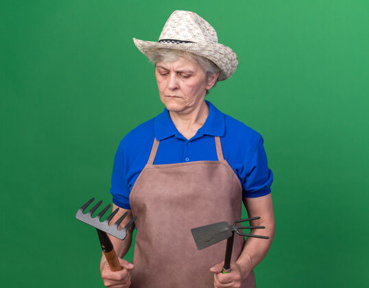帽子困惑的上了年纪的女园丁戴着园艺帽拿着耙子和锄头耙在草地上花园园艺困惑