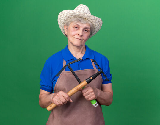 请高兴的上了年纪的女园丁戴着园艺帽拿着耙子和锄头耙在草地上老人女花园