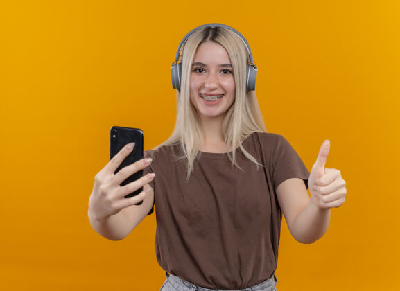 牙套微笑的金发女孩戴着耳机戴着牙套拿着手机 在孤立的橙色空间里竖起大拇指向上年轻抱着