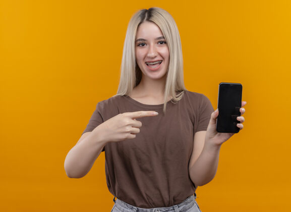 女孩快乐的金发女孩 戴着牙套 拿着手机 指着孤立的橙色空间和复印空间太空橙色持有