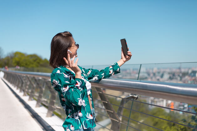 显示器阳光明媚的日子里穿着休闲绿衬衫的女人站在桥上看手机屏幕自拍视频通话无线蓝牙耳机在耳朵里青少年城市展示