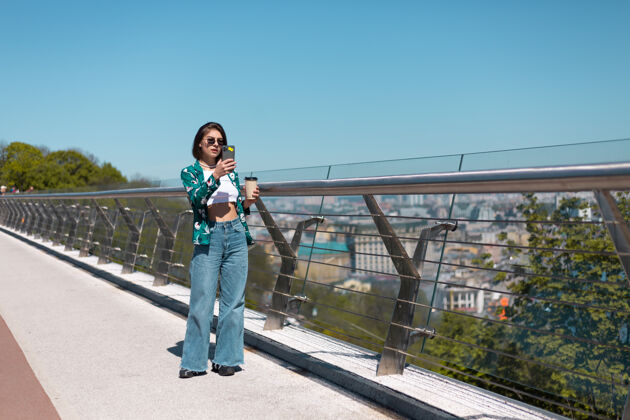 社交阳光明媚的日子里 穿着休闲绿衬衫和牛仔裤的女人在桥上散步 手机屏幕上放着一杯咖啡复制空间城市网络