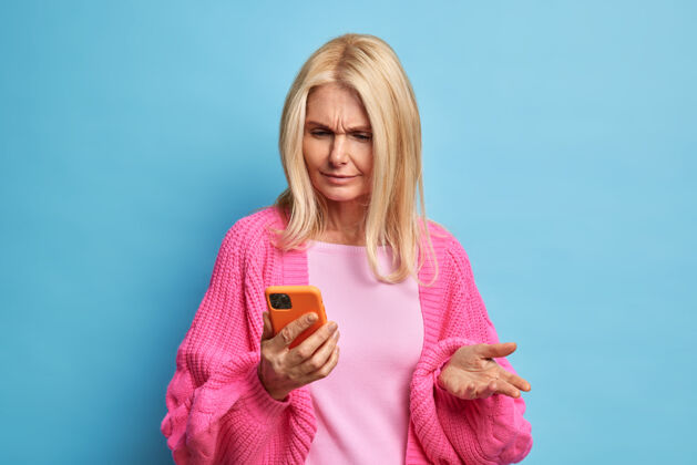 时尚迷惑不解的退休妇女用手机看着迷惑不解的笑脸 因为无法下载新的应用程序穿着休闲套头衫肖像金发傻笑