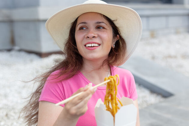 时尚年轻积极的女孩戴着帽子 用木筷吃中国面条作为外卖午餐的概念帽子魅力乌冬