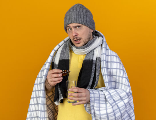 斯拉夫人无知的年轻金发病斯拉夫男子戴着冬季帽子和围巾包裹在格子布举行的药物在玻璃瓶的水玻璃隔离在橙色的墙壁与复制空间疾病药抱