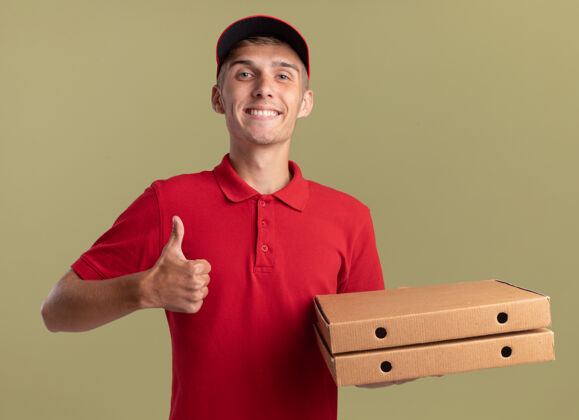 拿着年轻的金发送货员微笑着竖起大拇指 拿着橄榄绿的比萨饼盒金发披萨微笑