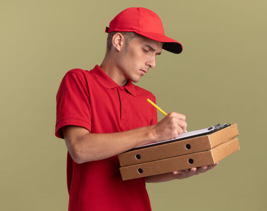 铅笔自信的年轻金发送货员用铅笔夹在橄榄绿的比萨饼盒上写在剪贴板上剪贴板披萨持有