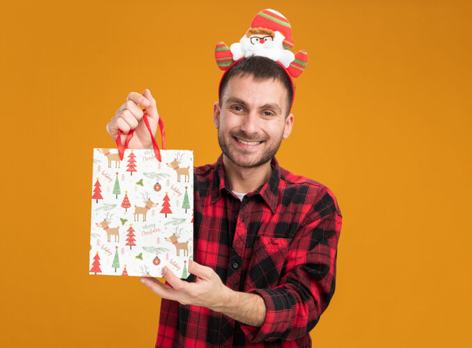 复制快乐的白人年轻人戴着圣诞老人的头带 手里拿着圣诞礼品袋 看着橙色背景上孤立的摄像机圣诞老人年轻空间