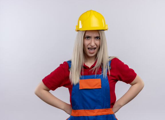 腰围愤怒的年轻金发工程师建筑女孩 穿着制服 戴着牙套 手放在腰上 孤立的白色空间愤怒金发年轻