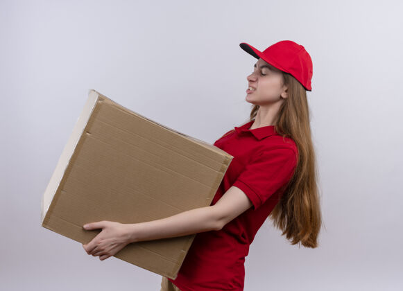 举行恼怒的年轻送货女孩穿着红色制服拿着箱子站在隔离的白色空间的侧视图交货红色女孩