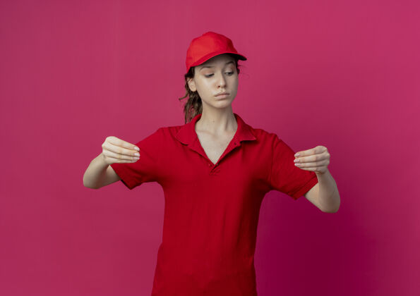 帽子自信的年轻漂亮的送货女孩穿着红色制服 戴着帽子 低头假装拿着深红背景上孤立的东西漂亮信心举行