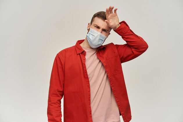 成人悲伤的年轻人留着胡子 穿着红衬衫 脸上戴着防毒面具 以防冠状病毒碰到他的额头 并在白墙上检查发烧年轻健康流感