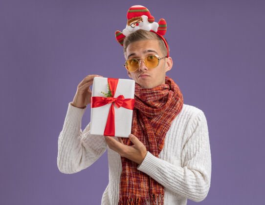 头带迷茫的年轻帅哥戴着圣诞老人的头带和围巾拿着礼包看着隔离在紫色背景上的相机举行围巾圣诞老人