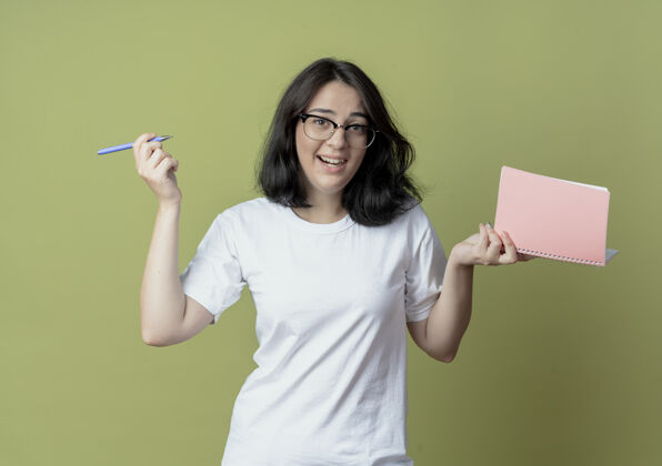 年轻令人印象深刻的年轻漂亮的白人女孩戴着眼镜拿着笔和笔记本孤立在橄榄绿的背景漂亮背景笔记
