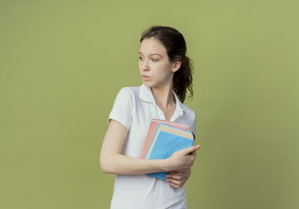 背景令人印象深刻的年轻漂亮的女学生拿着书和便笺簿看着一边孤立的橄榄绿背景与复印空间年轻垫女