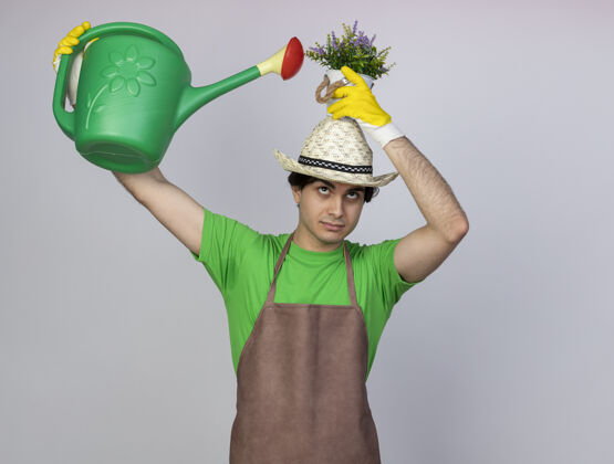 人高兴的年轻男园丁穿着制服 戴着园艺帽和手套 头上戴着浇花罐 在花盆里浇花花浇水花盆