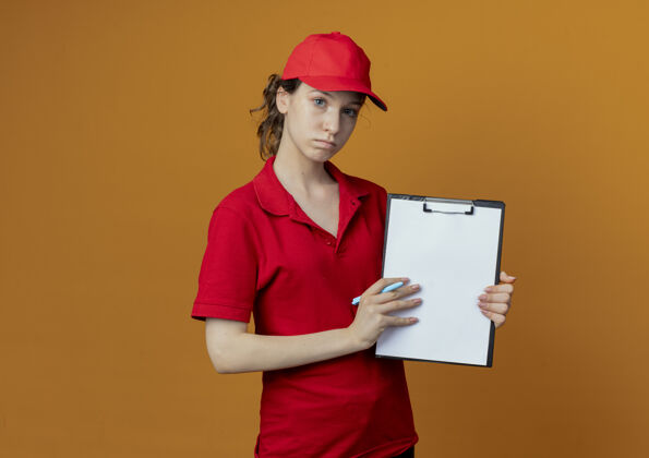 漂亮年轻漂亮的送货女孩 穿着红色制服 戴着帽子 看着相机 手里拿着笔 把剪贴板放在相机上 背景是橙色 有复印空间钢笔显示橙色