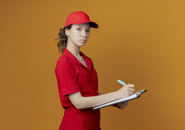 交货年轻漂亮的送货女孩穿着红色制服 戴着帽子站在侧视图中 手里拿着笔和剪贴板 准备写字 看着隔离在橙色背景上的相机 还有复印空间准备好了钢笔背景