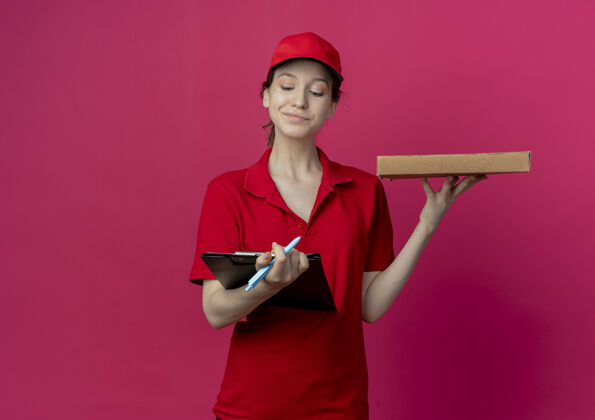 背景高兴的年轻漂亮的送货女孩在红色制服和帽子举行剪贴板和笔比萨饼包看着剪贴板上的深红色背景与复制空间隔离漂亮帽子制服