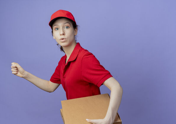 盒子令人印象深刻的年轻漂亮的送货女孩穿着红色制服 戴着帽子拿着披萨包 握紧拳头行走在紫色背景上 有复制空间送货漂亮背景