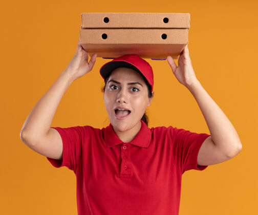 头快乐的年轻送货女孩穿着制服 戴着帽子 头上拿着披萨盒 隔离在橙色的墙上盒子年轻人脸