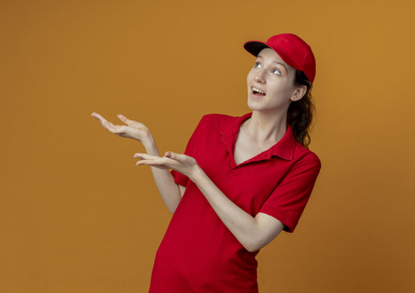 壁板令人印象深刻的年轻漂亮的送货女孩穿着红色制服 戴着帽子 看着和手指在一边孤立的橙色背景与复制空间漂亮女孩红色
