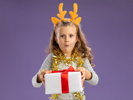 圣诞快乐兴奋的小女孩戴着圣诞发箍 脖子上戴着花环 拿着礼品盒站在蓝色背景的摄像机前兴奋抱着小