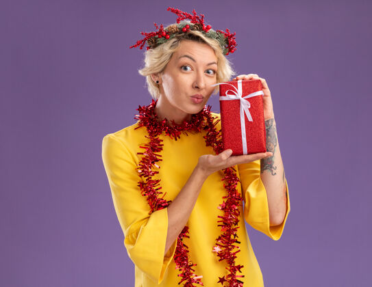 持有令人印象深刻的金发女郎戴着圣诞花环 脖子上戴着金属丝花环 手里拿着礼包 看着相机 噘起的嘴唇孤立在紫色的背景上紫色花环印象