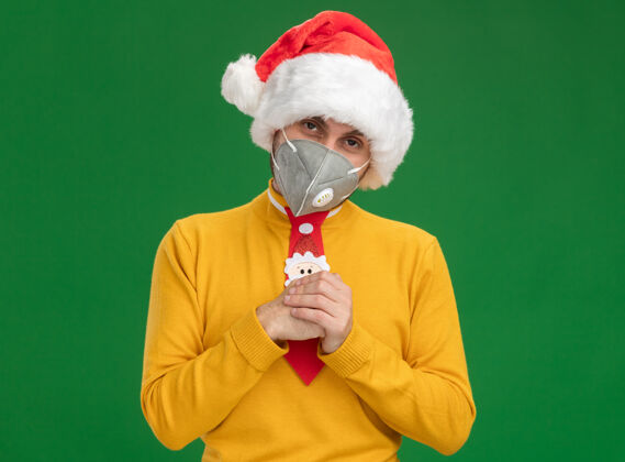 狡猾狡猾的白人年轻人戴着圣诞帽 打着带防护面具的领带 双手合十 看着绿色背景上的相机复制男人领带