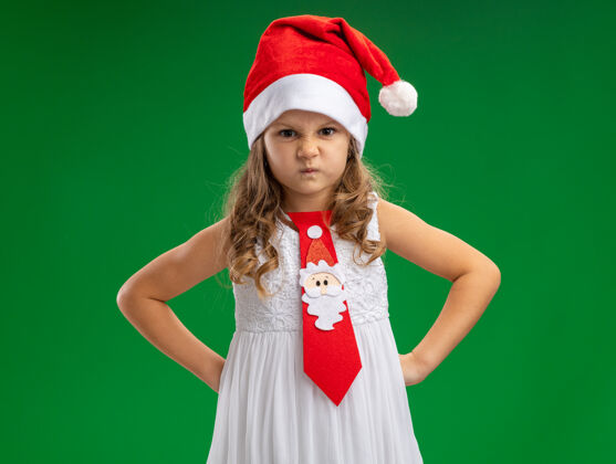 皱眉皱眉的小女孩戴着圣诞帽 打着领带 双手放在臀部 绿色背景上 与世隔绝圣诞节臀部圣诞快乐