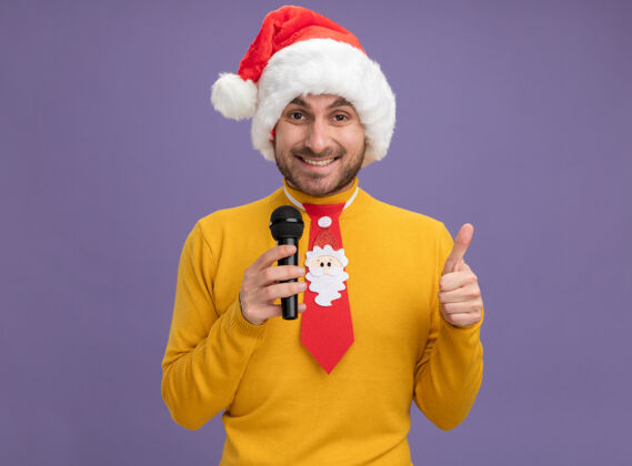 男人快乐的年轻白种人戴着圣诞帽打着领带拿着麦克风看着相机在紫色背景上孤立地竖起大拇指表演年轻向上