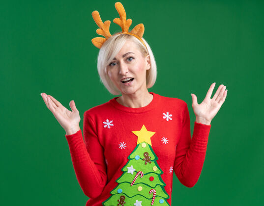 圣诞节令人印象深刻的中年金发女人穿着圣诞驯鹿鹿角头带和圣诞毛衣看着相机显示空手孤立的绿色背景中年鹿角毛衣