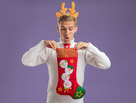 小伙子令人印象深刻的年轻帅哥戴着驯鹿鹿角头带和圣诞老人领带拿着圣诞长袜里面看紫色背景孤立驯鹿印象圣诞老人
