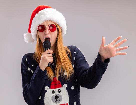 眼镜戴着圣诞帽戴着眼镜的快乐的年轻漂亮女孩在麦克风上唱歌 手被隔离在白色背景上白色圣诞快乐圣诞