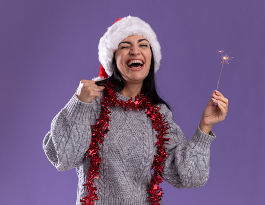 新年快乐的年轻白人女孩戴着圣诞帽 脖子上戴着金箔花环 手里拿着节日火花灯 做着“是”的手势 闭着眼睛 孤立地站在紫色的背景上是的金属片闪闪发光