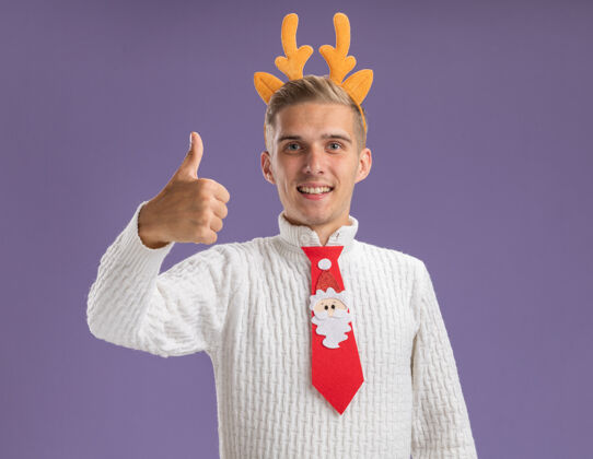 圣诞快乐快乐的年轻帅哥戴着驯鹿鹿角的头带和圣诞老人的领带看着相机 在紫色的背景上孤立地竖起大拇指驯鹿年轻头带