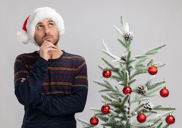 帽子令人印象深刻的年轻白人男子戴着圣诞帽站在圣诞树旁触摸下巴看着孤立的白色背景圣诞快乐印象触摸