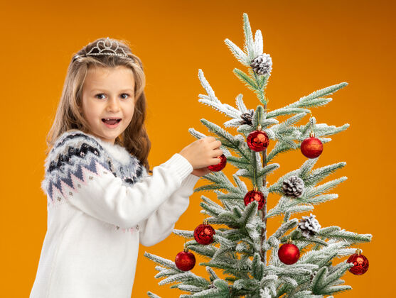 站快乐的小女孩站在圣诞树旁边 戴着头饰 脖子上戴着花环 把树孤立在橙色的背景上快乐小手拿