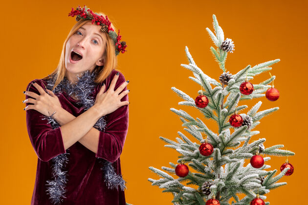 橙色兴奋的年轻漂亮的女孩站在圣诞树旁边 穿着红色的裙子 脖子上戴着花环 手放在肩膀上 橙色的背景孤立着手花环脖子