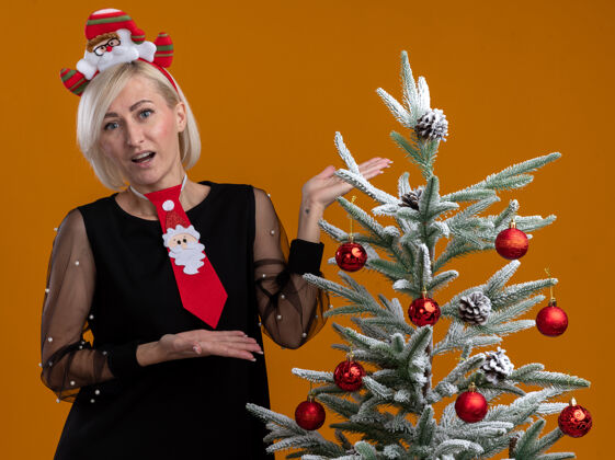 站令人印象深刻的中年金发女人戴着圣诞老人的头带和领带站在装饰圣诞树附近 用手看着橙色背景上孤立的摄像头圣诞中年圣诞老人
