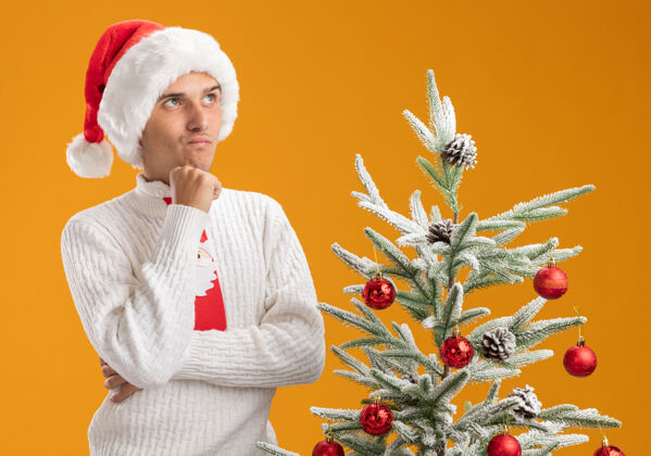橙色戴着圣诞帽 系着圣诞老人领带的年轻帅哥抱着怀疑的态度站在装饰好的圣诞树旁 孤零零地仰望着橙色的背景年轻领带圣诞快乐