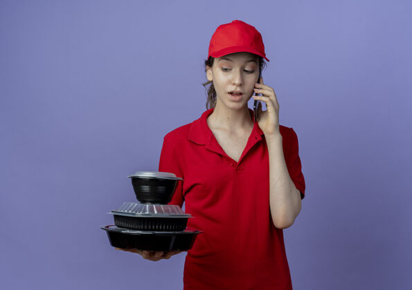 年轻令人印象深刻的年轻漂亮的送货女孩穿着红色制服 戴着帽子 看着食品容器 在紫色背景上与复印空间隔离的电话交谈背景容器印象