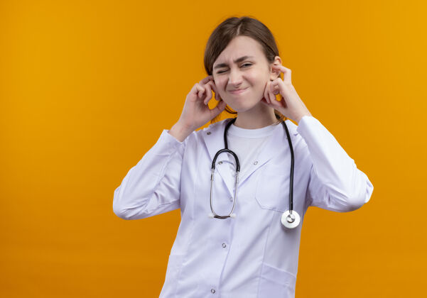女恼怒的年轻女医生穿着医用长袍和听诊器把手放在耳朵上孤立的橙色空间与复制空间穿年轻空间