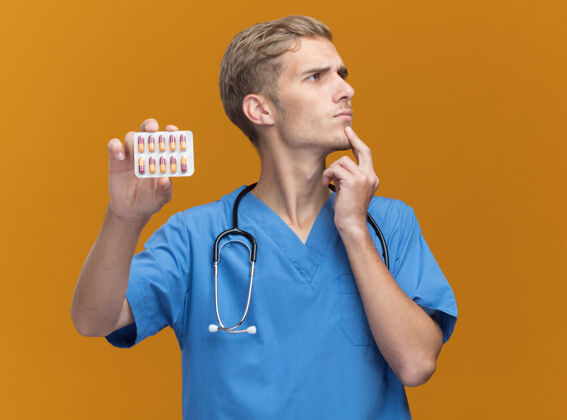 姿势想着看着身边年轻的男医生穿着医生制服拿着听诊器拿着药片把手放在下巴下面隔离在橘色的墙上思维衣服男性