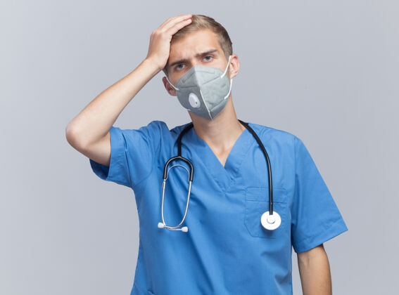 遗憾遗憾的是 年轻的男医生穿着医生制服 戴着听诊器和医用口罩 手放在隔离在白墙上的头上表情男性面具