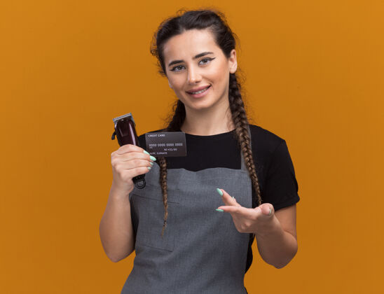 制服年轻的女理发师面带微笑 拿着制服 指着隔离在橙色墙上的信用卡和理发器脸人剪子