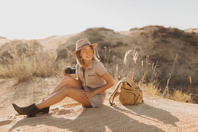 相机沙漠中穿着卡其色长裙的迷人时尚年轻女子 在非洲旅行 戴着帽子和背包 用老式相机拍照体验旅行时尚