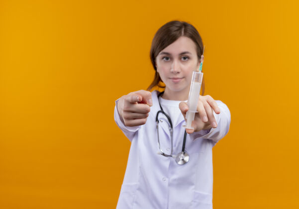 穿自信的年轻女医生穿着医用长袍 手持注射器 手持听诊器 指着隔离的橙色空间和复印空间医疗听诊器注射器